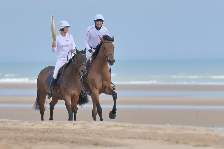 la cavalière Pénélope Leprévost (g) porte la flamme olympique, accompagnée de l'humoriste Nicolas Canteloup, sur la plage d'Omaha Beach, le 30 mai 2024 à Saint-Laurent-sur-Mer, dans le Calvados