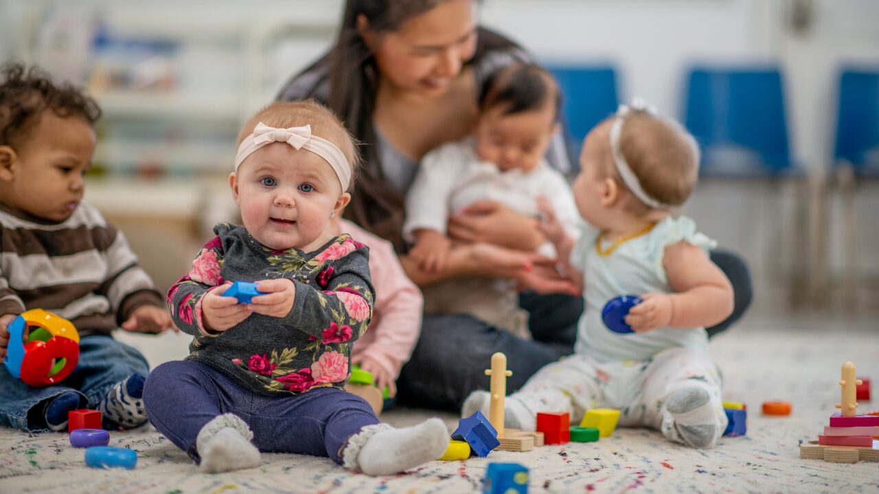 Une auxiliaire de puériculture s'occupe de 4 bébés. Au premier plan, un des enfants fixe l'objectif de la caméra.