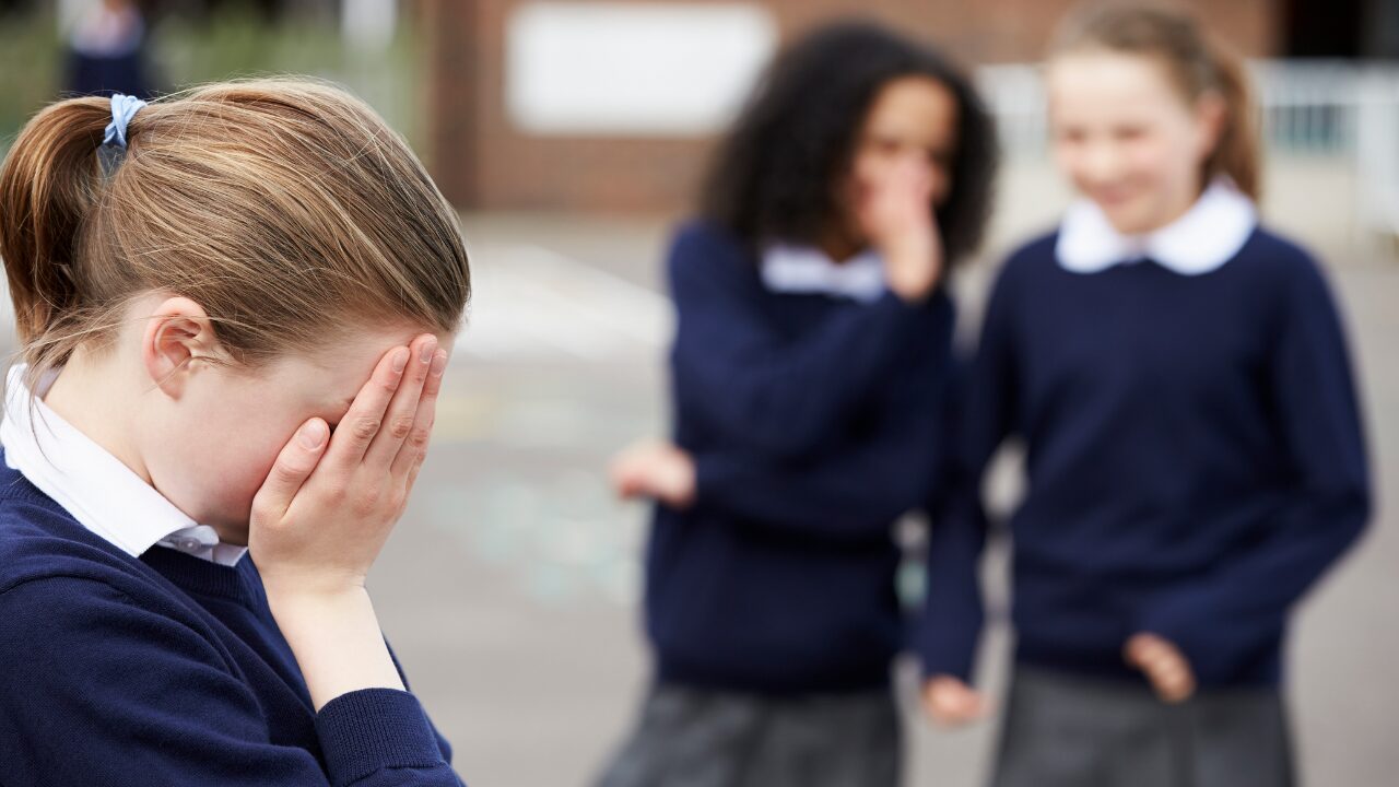 Dans une cour de récréation, une élève se cache les yeux, à l'écart de deux autres jeunes filles qui se moquent d'elles. La jeune fille isolée semble victime de harcèlement scolaire