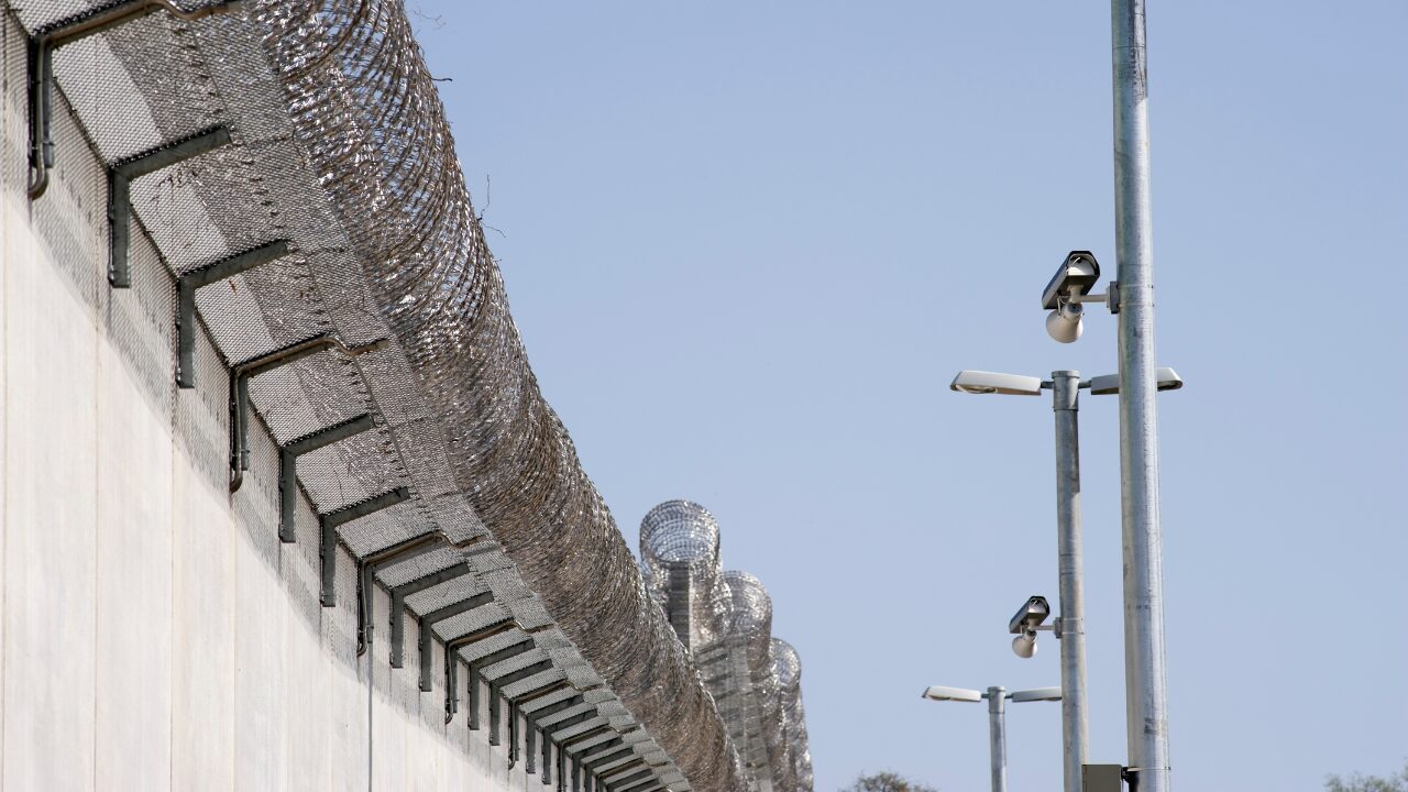 La photo représente le mur entourant une prison. Une longue rangée de barbelés encadre la cloison qui sépare les prisonniers de l'extérieur. À droite de l'image, des caméras surveillent la zone